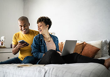 Man and woman sitting on a bed, looking at their electronics